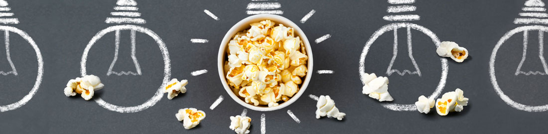 Chalk drawn lightbulbs on a gray background and in the middle of the middle bulb is a bowl of popcorn with kernels laid about.