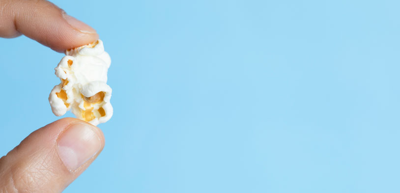 close up shot of fingers hold a single piece of white popcorn on a baby blue background