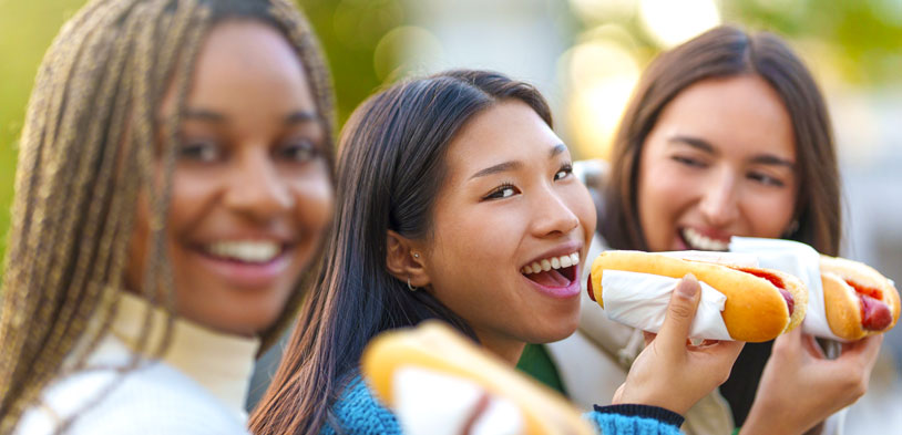 girls next to each other eating hot dogs in the park