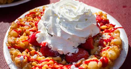 Funnel cake with Whip Cream and Strawberries on top with a strawberry sauce drizzeled over.