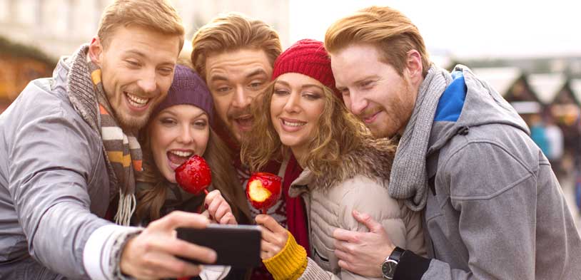 Group of friends bundled in winter clothes taking a selfie while enjoying red candy apples at an outdoor event.