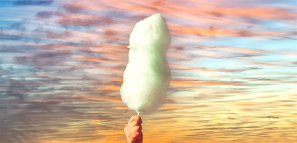 A hand holds cotton candy in front of clouds on a sunset backdrop