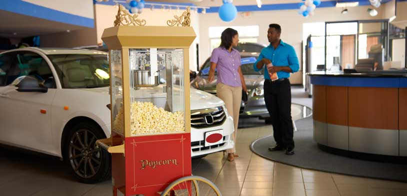 Sales person and customer talk in a car dealership while the sales person eats from a bag of popcorn the dealership popped with their popcorn machine.