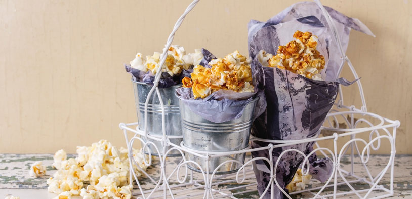 Gourmet popcorn in bucket tins and paper cones sitting in a white wire tray. Gourmet popcorn in bucket tins and paper cones sitting in a white wire tray.