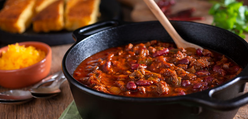 A pot of chili in cast iron pot on table with cheese and bread next to it.