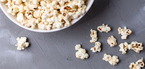 A bowl of white popcorn sit with a gray backdrop and pieces of popcorn strewn about