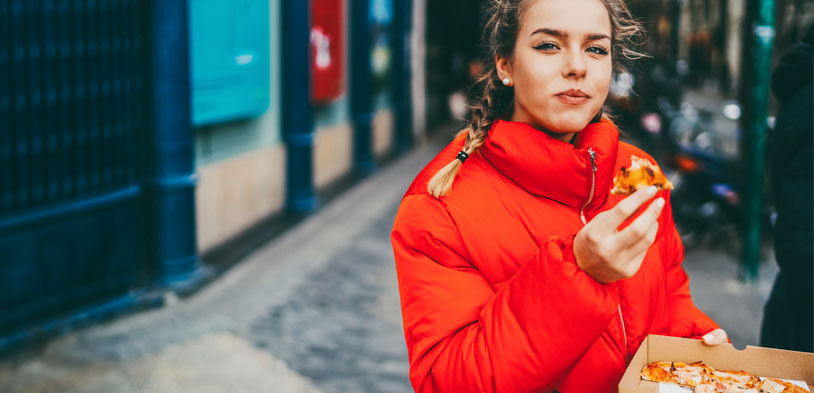 woman eats pizza on street while wearing orange puffy coat