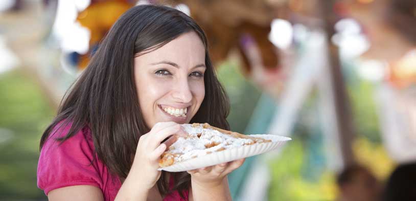 Smiling woman in a pink shirt enjoying a powdered sugar funnel cake at an outdoor fair.