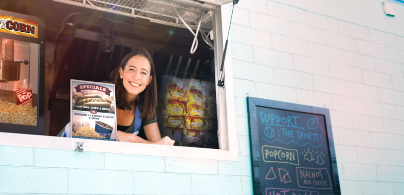 A women in a concession stand leans on counter smiling surrounded by ready to eat snacks and fresh popped popcorn.
