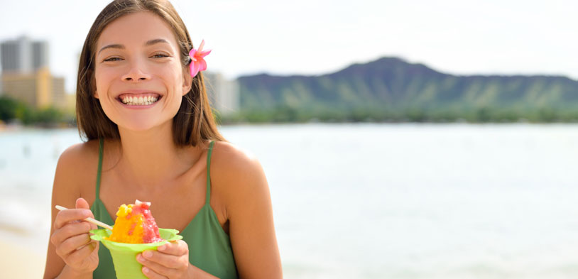 a smiling woman in front of a ocean side background enjoys shaved ice.