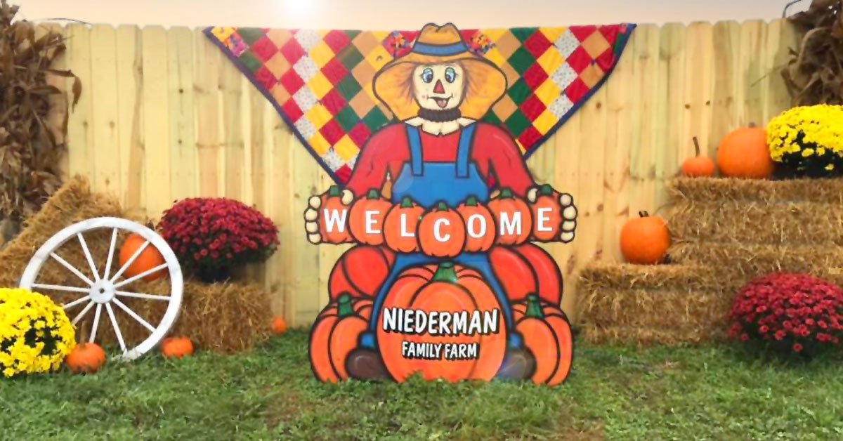 wooden scarecrow figure holding welcome to Niederman Family Farm sign with a picket fence and autumn background