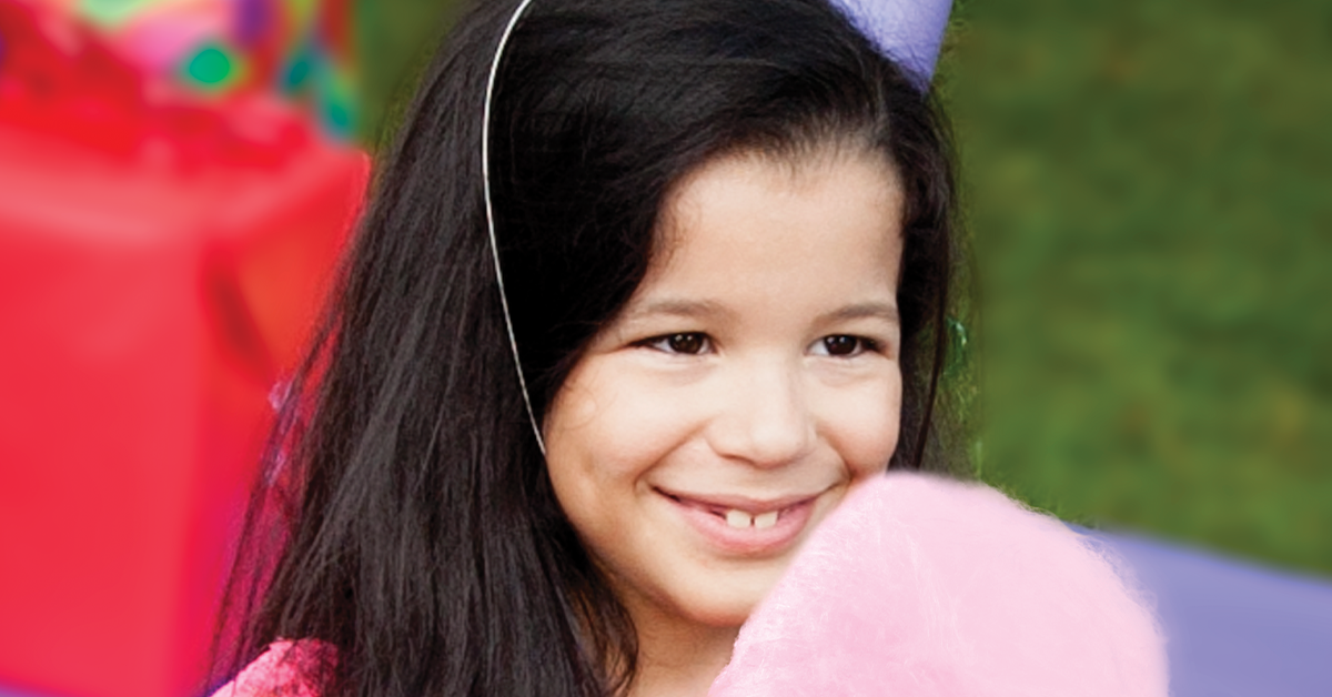 young girl wearing a party hat holding pink cotton candy with gifts and a green lawn blurred in background