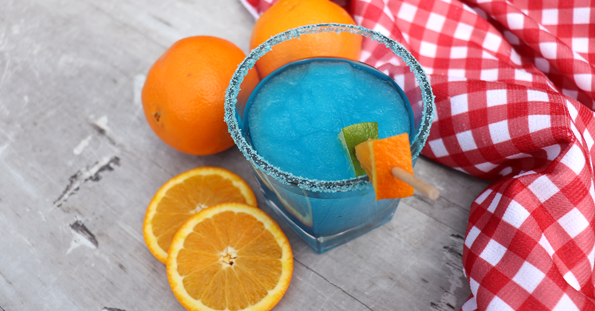 blue ocean ice slush in glass lined with piña colada flossugar sugar garnished with lime and orange on a red and white checkered cloth surrounded by oranges