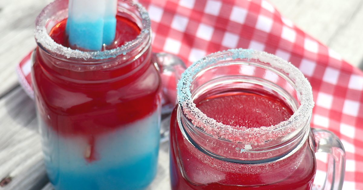 red, white and blue patriotic pop frozen slush drink in a glass with popsicles sitting on a checkered red and white cloth