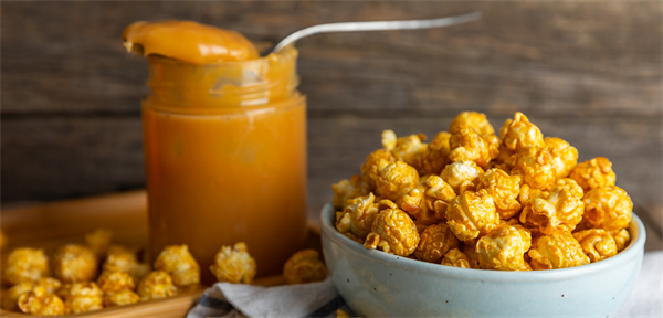 Close-up of gourmet caramel popcorn in a light blue bowl next to a jar of rich, creamy caramel with a spoon, set against a rustic wooden background.