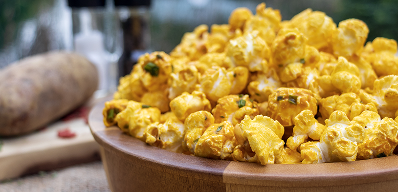Bowl of savory yellow loaded baked poptato popcorn seasoned with green herbs, styled on a rustic wooden surface with a whole potato and salt shaker in the background. Bowl of savory yellow loaded baked poptato popcorn seasoned with green herbs, styled on a rustic wooden surface with a whole potato and salt shaker in the background.