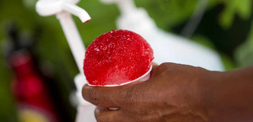A man holds a snow cone in his hand and pumps syrup onto it.