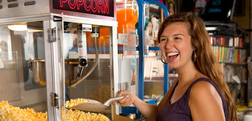 A woman smiles as she scoops popcorn out of a popcorn machine by gold medal products with new LED Red and Black Popcorn sign.